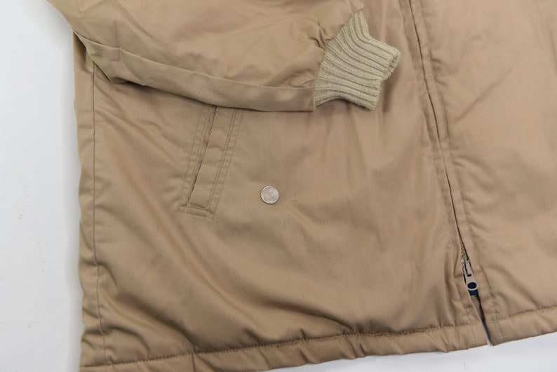 May include: A tan jacket with a zipper closure and a ribbed knit cuff. The jacket has a pocket on the left side and a zipper pull with a blue tab.