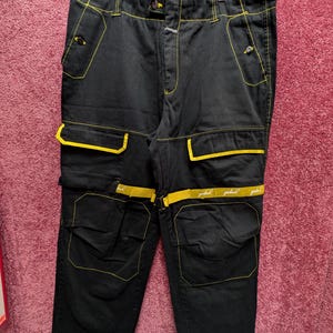 May include: Black cargo trousers with yellow stitching and detailing. The trousers have multiple pockets, yellow trim, and a button closure. The yellow stitching outlines the pockets and seams, creating a contrasting design. The trousers are hanging on a wooden hanger.