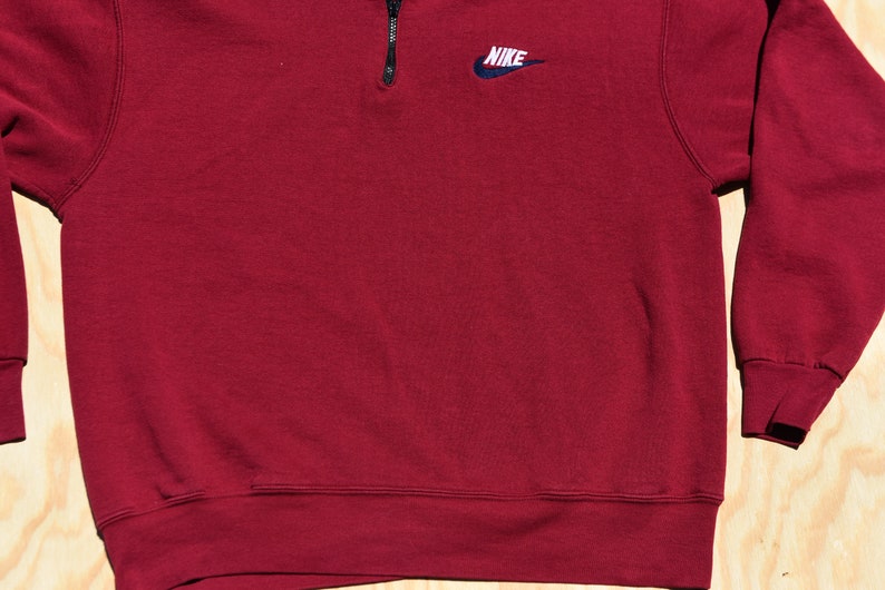 May include: A maroon 1/4 zip sweatshirt with a small embroidered Nike logo on the left chest.