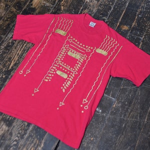 May include: A red t-shirt with a gold geometric design. The design features lines, dots, and triangles.