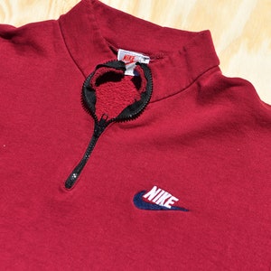 May include: A red quarter-zip sweatshirt with a black zipper and a navy blue Nike logo on the front. The logo has the word "NIKE" in white lettering.