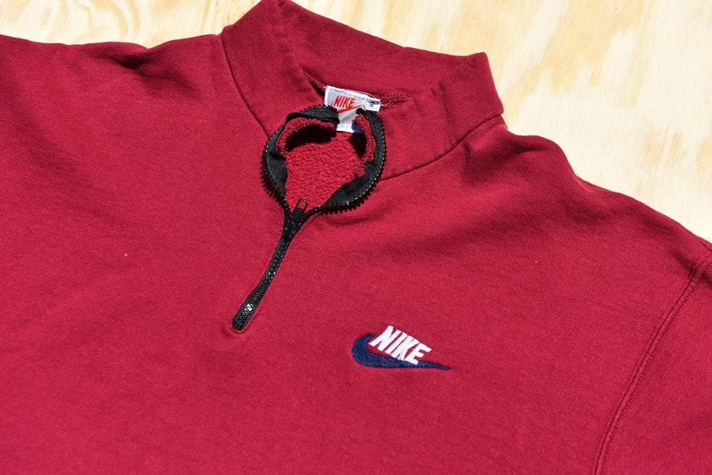 May include: A red quarter-zip sweatshirt with a black zipper and a navy blue Nike logo on the front. The logo has the word "NIKE" in white lettering.
