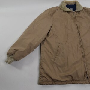 May include: A tan zip-up jacket with a ribbed collar and cuffs. The jacket has a zipper closure and two pockets on the front.