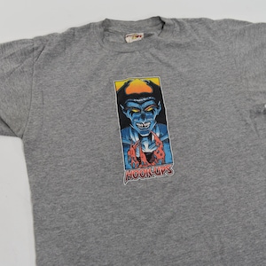 Puede incluir: Camiseta gris con un estampado gráfico de un demonio azul con detalles rojos y amarillos. El gráfico incluye el texto "Hook-Ups".