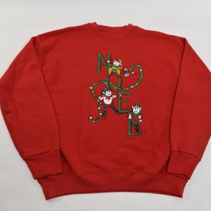 以下が含まれることがあります： 「NOEL」と綴られたライトの紐を登る3人のエルフが描かれた、赤いスウェットシャツです。