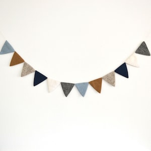 May include: A felt banner with triangular flags in shades of blue, brown, and white. The banner is strung on a white wall.