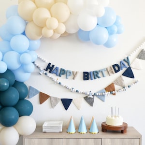 May include: A festive birthday party scene with a balloon arch in shades of blue and cream. A "HAPPY BIRTHDAY" banner hangs above a garland of felt pennants. Party hats, a cake with candles, and plates are arranged on a wooden cabinet.