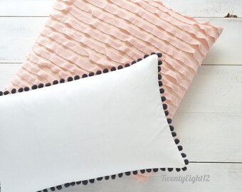 May include: Two decorative throw pillows with a white cotton pillow with a black pom-pom trim on top of a pink ruffled pillow with a black pom-pom trim.