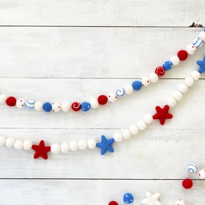 May include: A red, white, and blue felt garland with stars and polka dots. The garland is strung on white felt balls. The garland is hanging on a white wooden background.