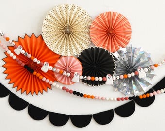 May include: Halloween-themed decorations featuring orange, pink, black, and silver paper fans and felt ball garlands. The garlands include small white ghost figures. A black scalloped banner is at the bottom, creating a festive display.