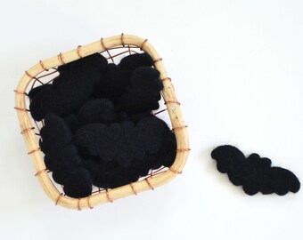 May include: A square woven basket filled with black felt bat shapes. One bat shape is outside the basket. The basket has a light brown wooden frame and copper wire accents. The bats are a solid black color.
