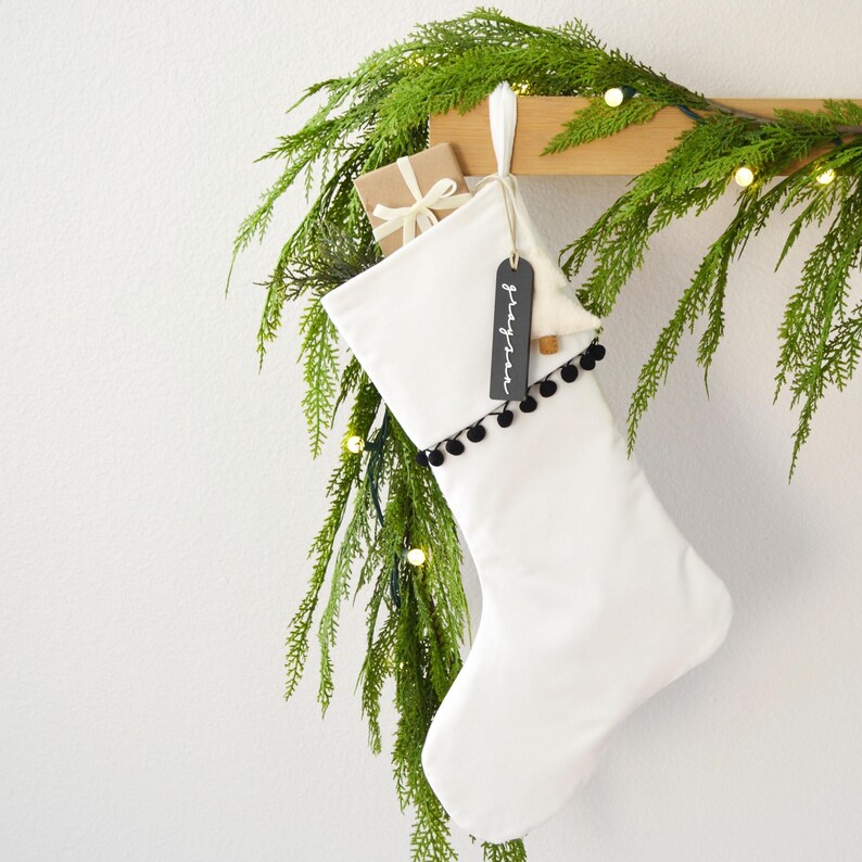 A white Christmas stocking adorned with greenery and a black tag hangs on a wooden hanger, with a string of lights visible in the background.