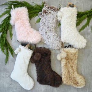 May include: Six fluffy Christmas stockings in various colors: light pink, gray, white, dark brown, and tan. Each stocking has a loop for hanging and is made of a soft, textured material. Greenery is arranged above the stockings.