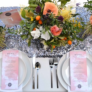 May include: A wedding table setting with two place settings, each with a white plate, a white napkin, a silver fork and knife, a silver spoon, and a pink menu card with the text "MENU" and "Olivia + Holden". The table is covered with a silver sequin tablecloth and decorated with floral centerpieces.