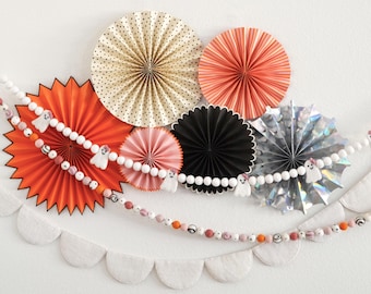 May include: A Halloween-themed wall decoration featuring orange, black, pink, and silver paper fans. A garland of white beads with ghost figures and a scalloped banner complete the festive display. The fans are arranged in a circular pattern.