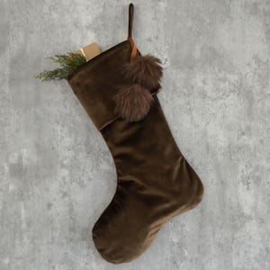 May include: Brown velvet Christmas stocking with a loop for hanging. The stocking is decorated with two brown pom-poms and is filled with a small gift and greenery. The stocking is approximately 18 inches long.