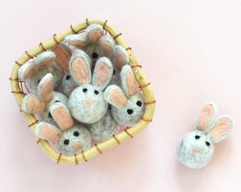May include: A wicker basket filled with gray felt bunny ornaments. One bunny is sitting outside the basket on a pink surface.