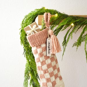 May include: A knitted Christmas stocking with a pink and white checkerboard pattern, a pink cuff, and a matching tassel. A small gift wrapped in brown paper and tied with a white ribbon is inside. A wooden tag reads "Sophie".