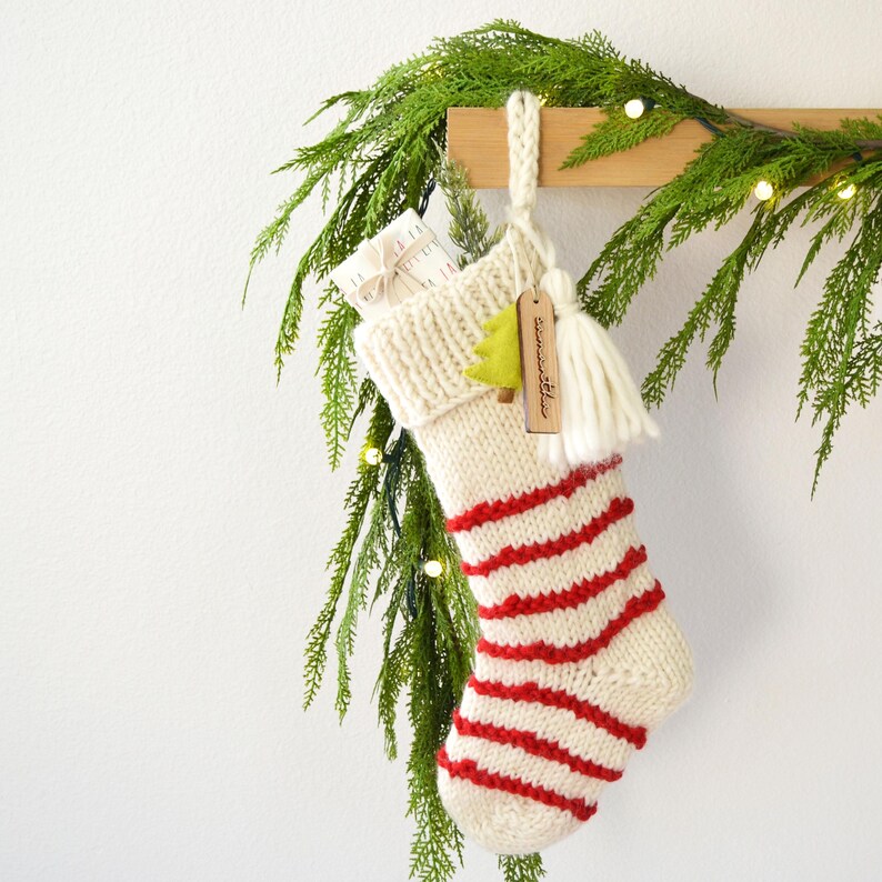 A white and red striped Christmas stocking with a tassel hangs on a wooden hanger, decorated with greenery and twinkling lights.