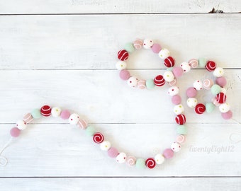 May include: A festive garland made of felt balls in pink, red, white, and green with white polka dots, swirls, and stripes. The garland is strung together with white thread and is perfect for decorating for the holidays.