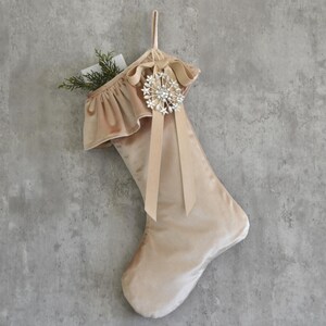 May include: A champagne-colored satin Christmas stocking featuring a ruffled cuff, a hanging loop, and a decorative silver-toned starburst brooch with clear crystals. A small sprig of green pine needles is tucked into the top of the stocking.