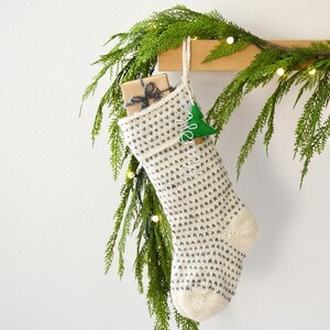 A white and gray patterned stocking hangs from a wooden hanger, adorned with greenery and twinkling lights, creating a festive and cozy holiday scene.