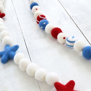 May include: A white, blue, and red felt ball garland with stars and polka dots. The garland is strung on a white background.