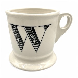 以下が含まれることがあります： 前面に黒い文字「W」が付いたクリーム色のセラミックマグ。文字は花柄とストライプ柄で装飾されています。マグにはハンドルと台座が付いています。