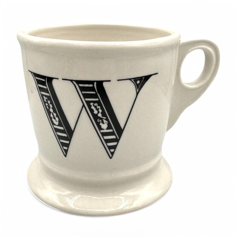 以下が含まれることがあります： 前面に黒い文字「W」が付いたクリーム色のセラミックマグ。文字は花柄とストライプ柄で装飾されています。マグにはハンドルと台座が付いています。