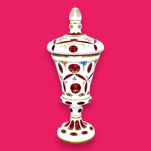 Puede incluir: Un tarro decorativo blanco y rojo con tapa y detalles dorados. El tarro tiene una base de pedestal y una tapa redondeada. El cuerpo está decorado con diseños circulares rojos y motivos florales. El fondo es rosa liso.