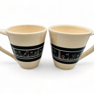 Può includere: Due tazze in ceramica color crema con bande nere e un motivo floreale. Le tazze hanno una forma arrotondata e un manico.