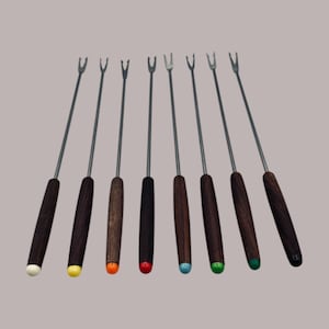 Può includere: Un set di otto forchette da fonduta con manici in legno scuro e rebbi in acciaio inossidabile. I manici hanno punte colorate in bianco, giallo, arancione, rosso, blu e verde. Sfondo neutro.