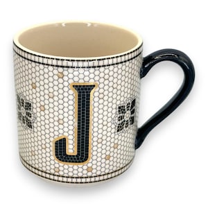 Op de afbeelding: Witte keramische mok met een zwarte handgreep en de letter "J" in zwart-gouden mozaïektegels. De mok heeft een honingraatpatroon en decoratieve bloemelementen. De binnenkant is beige.