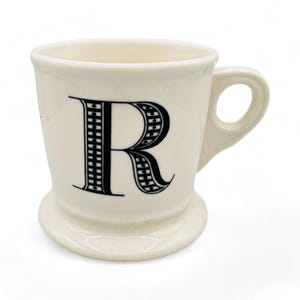 Può includere: Una tazza in ceramica bianca con una lettera "R" a motivi bianco e nero sul davanti. La tazza ha una base larga e un manico.