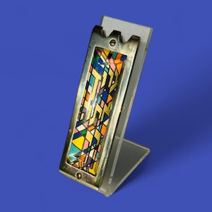 May include: A silver-toned mezuzah case with a colorful stained-glass design. The case is rectangular with a decorative top and is mounted on a clear acrylic stand. The stained glass features geometric shapes in various colors, including blue, yellow, and orange.