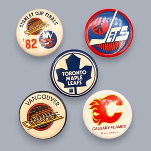 May include: A collection of vintage hockey pins featuring team logos and text. The pins include the Vancouver Canucks, Toronto Maple Leafs, Calgary Flames, and Winnipeg Jets. The pins are round with various colors and designs.