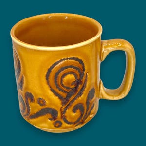 Può includere: Una tazza in ceramica con una glassa marrone dorato e un motivo a spirale marrone scuro. La tazza ha un manico curvo e una finitura liscia e lucida. Anche l'interno della tazza è marrone dorato.