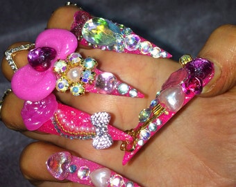 May include: Pink and white long acrylic nails with rhinestones and glitter. The nails are decorated with a pink flower, a bow, and a heart.