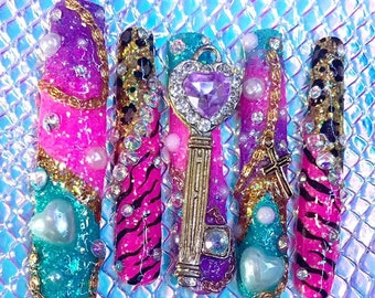 May include: Set of five colorful fake nails with various designs, including a pink and black zebra print, a purple and gold design with a key charm, and a turquoise and gold design with a cross charm.