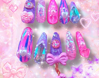 May include: Set of 10 colorful, glittery, and decorated fake nails with a pink bow charm. The nails are in various shades of pink, purple, and blue with glitter and pearl accents.