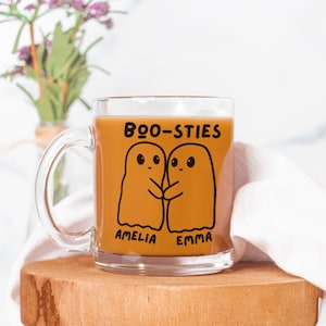 May include: A clear glass mug filled with a light brown liquid, featuring a black graphic of two hugging ghosts with the text "BOO-STIES" above them. Below the ghosts are the names "AMELIA" and "EMMA". The mug sits on a wooden surface.