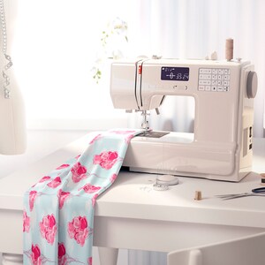 May include: A white sewing machine with a digital display showing "0-35.24". The machine is on a white table with a piece of fabric with pink flowers on it. There are spools of thread, scissors, and other sewing supplies on the table.