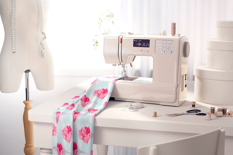 May include: A white sewing machine with a digital display showing "0-35.24". The machine is on a white table with a piece of fabric with pink flowers on it. There are spools of thread, scissors, and other sewing supplies on the table.