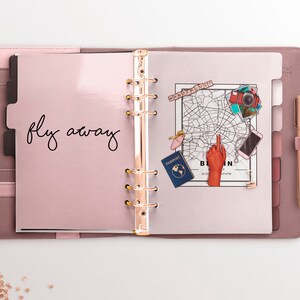 May include: A pink leather bound planner with gold rings and a white page open. The page has the words "fly away" written in black ink. There are stickers on the page, including a map of Berlin, a passport, a camera, and a hand pointing to the map.