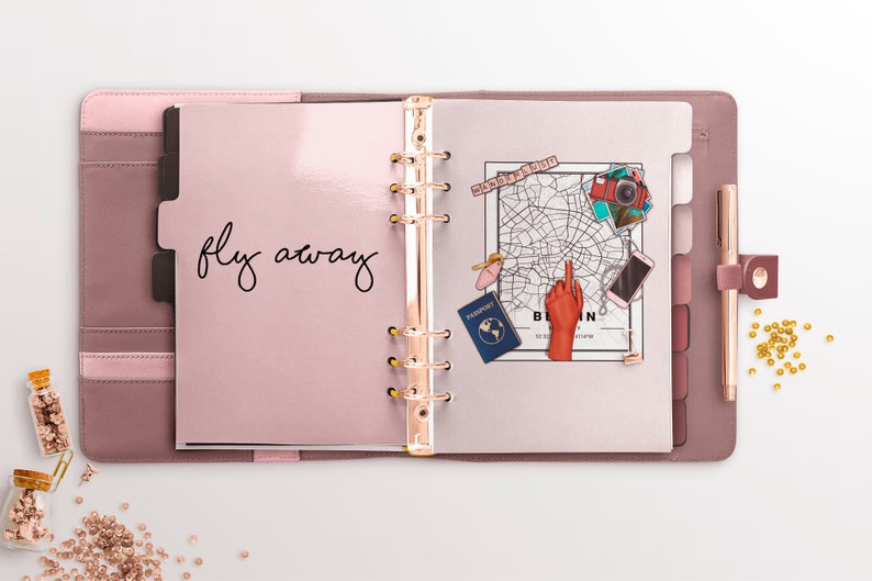 May include: A pink leather bound planner with gold rings and a white page open. The page has the words "fly away" written in black ink. There are stickers on the page, including a map of Berlin, a passport, a camera, and a hand pointing to the map.