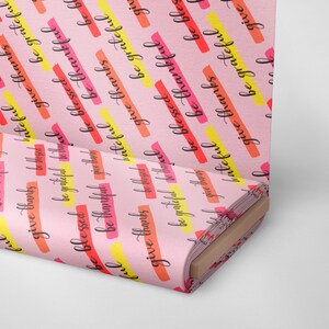 Op de afbeelding: Roze stof met een herhalend patroon van de woorden "give thanks", "be blessed", "be grateful" in zwarte letters. De woorden zijn gelegd over kleurrijke strepen in geel, oranje en rood.