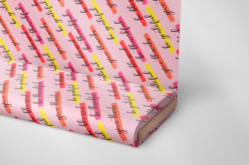 Op de afbeelding: Roze stof met een herhalend patroon van de woorden "give thanks", "be blessed", "be grateful" in zwarte letters. De woorden zijn gelegd over kleurrijke strepen in geel, oranje en rood.
