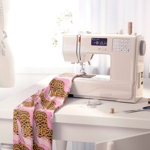 May include: A white sewing machine with a digital display showing "0-0 35.24". The machine is on a white table with a pink and brown leopard print fabric, spools of thread, and a pair of scissors.