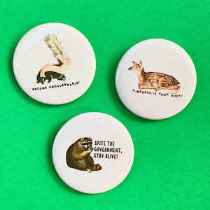May include: Three white round buttons with animal illustrations and text. One button features a skunk with the text "BECOME UNGOVERNABLE!". Another shows a deer with "KINDNESS IS PUNK ROCK!". The last button has a raccoon and reads "SPITE THE GOVERNMENT, STAY ALIVE!"