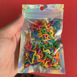 May include: A clear, iridescent bag filled with colorful, squiggly sprinkles. The sprinkles are in various colors, including pink, orange, yellow, green, and blue. The bag has the text "1 oz" printed on it.